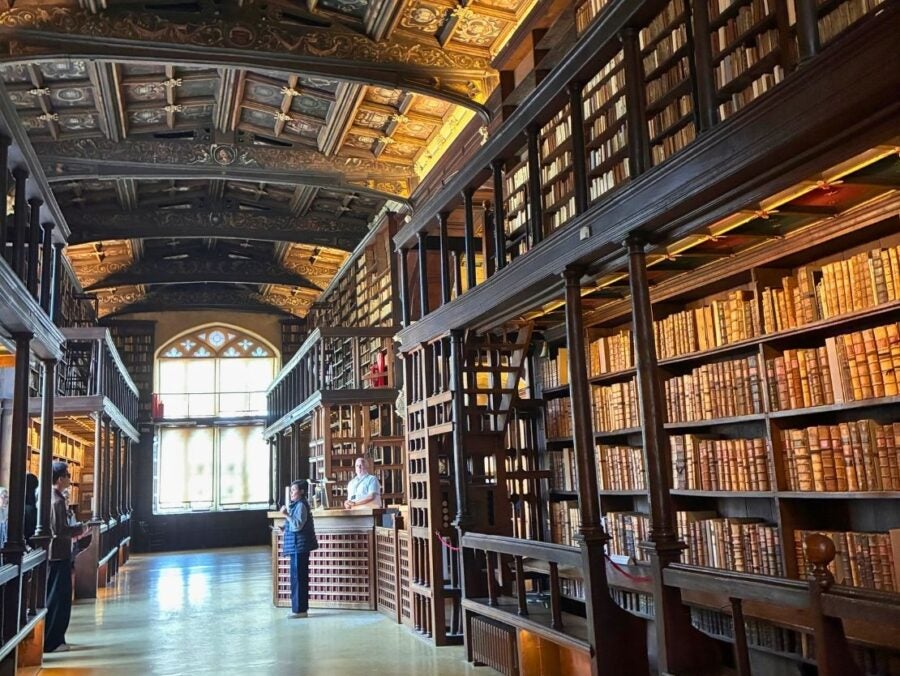 Historic library interior with tall wooden bookshelves filled with old leather-bound books and a decorative vaulted ceiling.