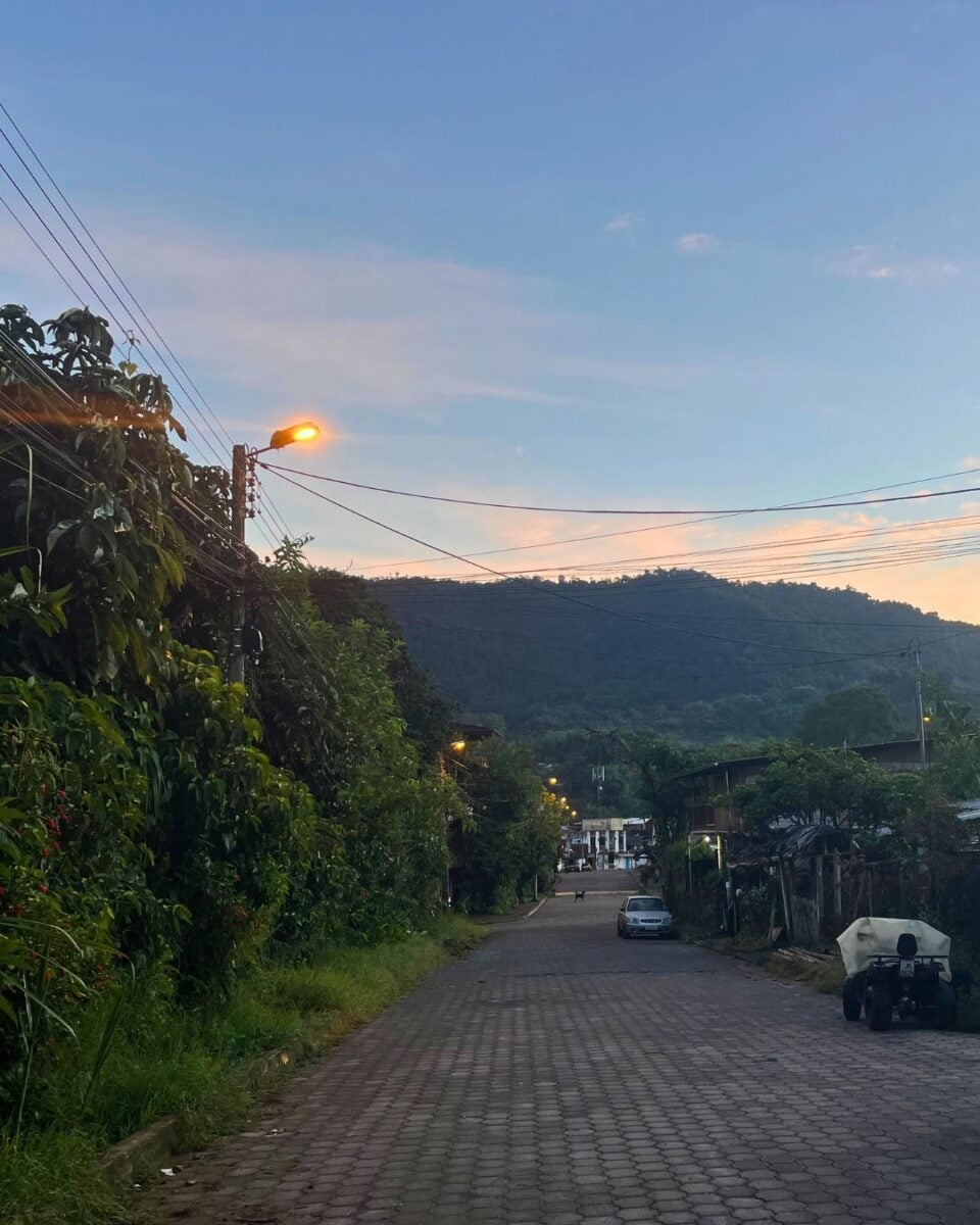 Quiet cobblestone street at dusk with streetlights glowing and hills in the background under a clear sky.