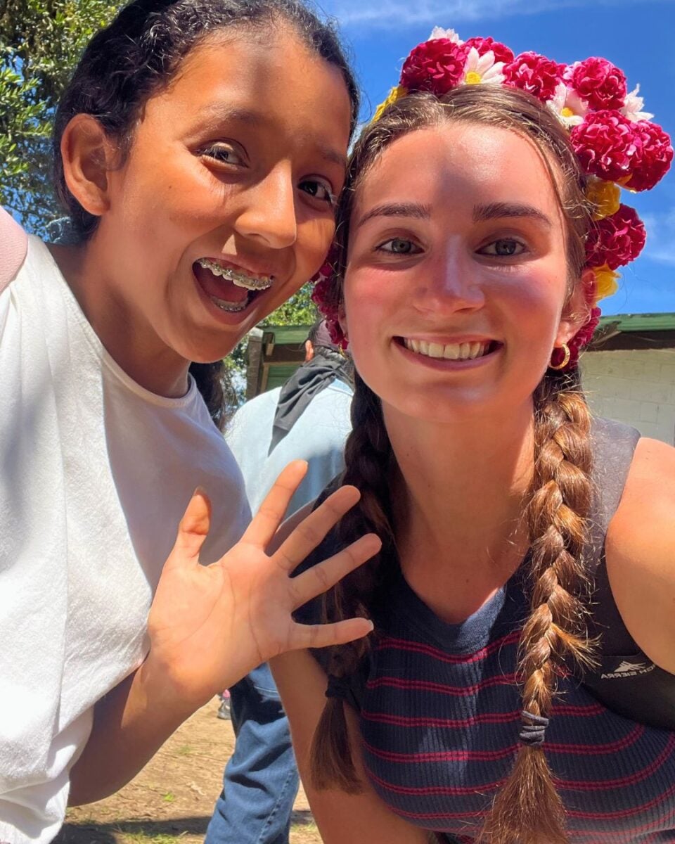Two young women smiling outdoors, one with braces waving and the other wearing a colorful flower crown and braided hair.