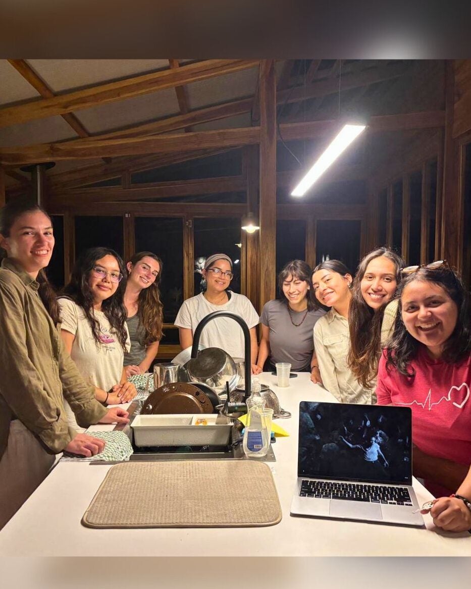 Group of eight women gathered around a kitchen island smiling and watching a laptop screen together at night.