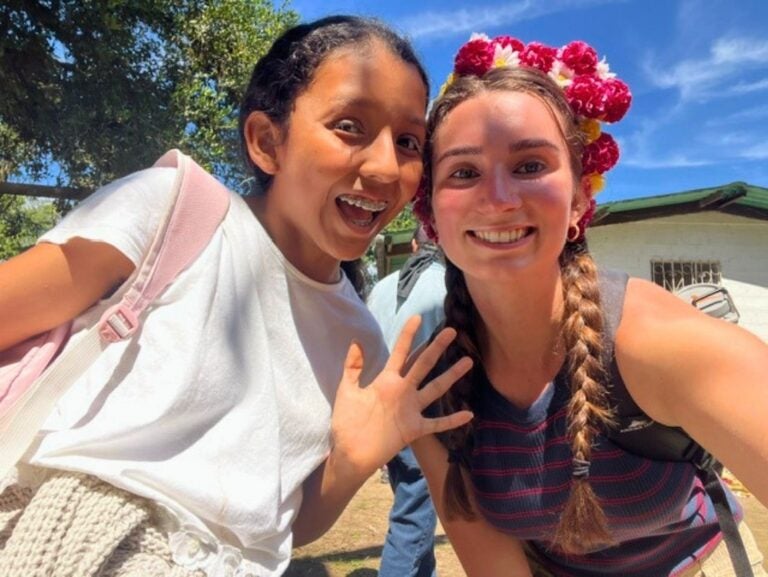 Two young women smiling outdoors, one with braces waving and the other wearing a colorful flower crown and braided hair.
