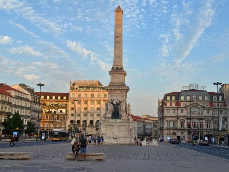 Monumento aos Restauradores. Tall stone obelisk monument with statues at its base in a bustling city square during sunset.