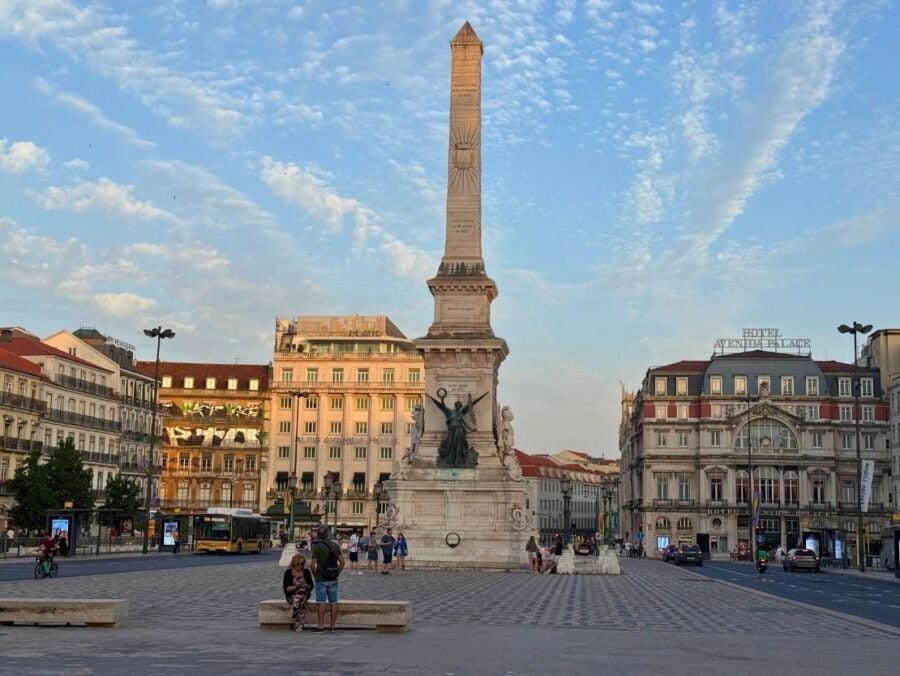 Monumento aos Restauradores. Tall stone obelisk monument with statues at its base in a bustling city square during sunset.