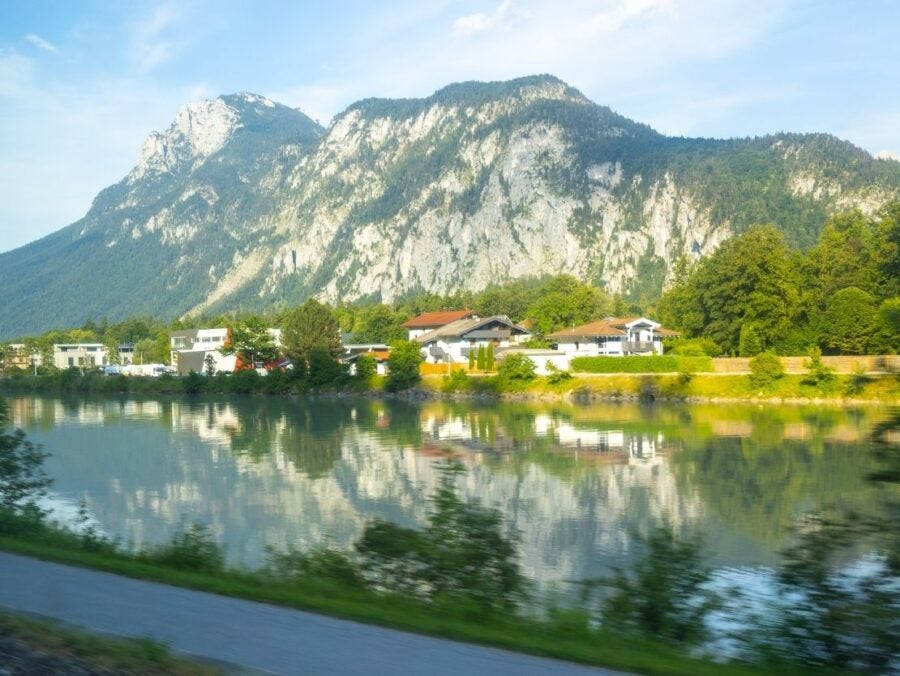 Scenic view of a mountain range reflected in a calm river with houses and greenery along the shore under a blue sky.