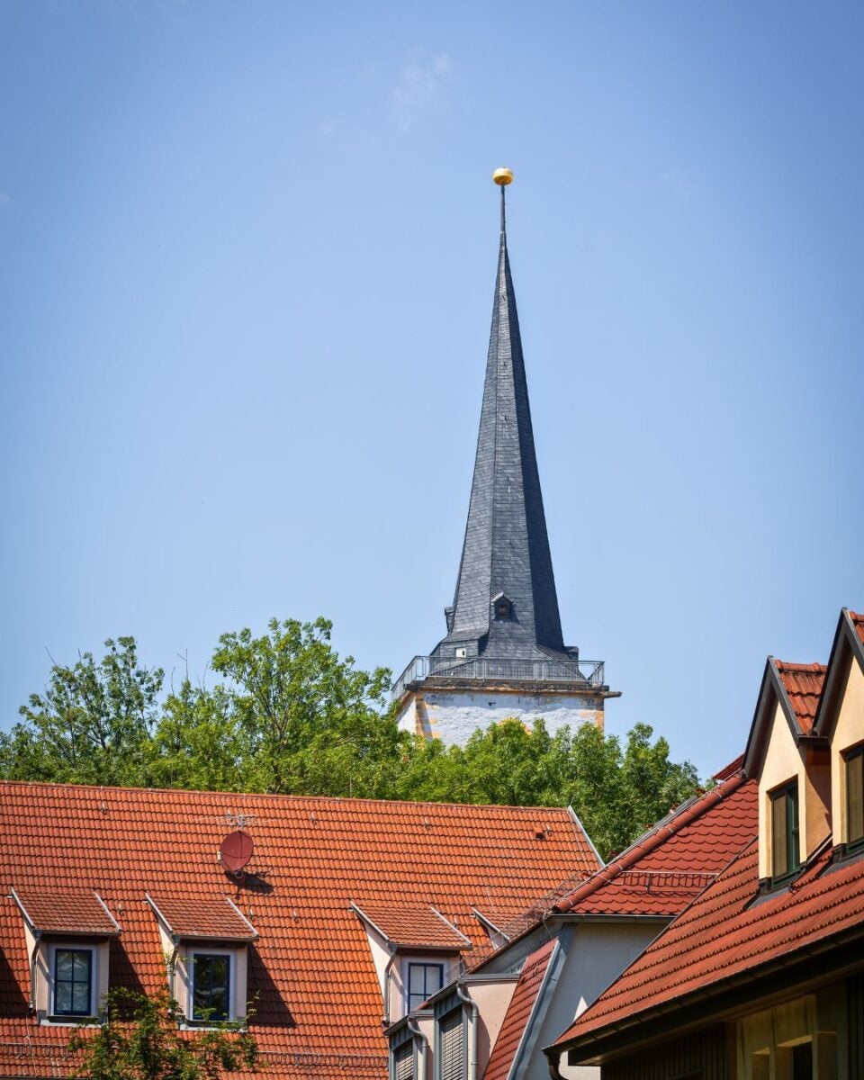 Tall church steeple with a golden sphere on top rising above green trees and red-tiled rooftops under a clear blue sky.