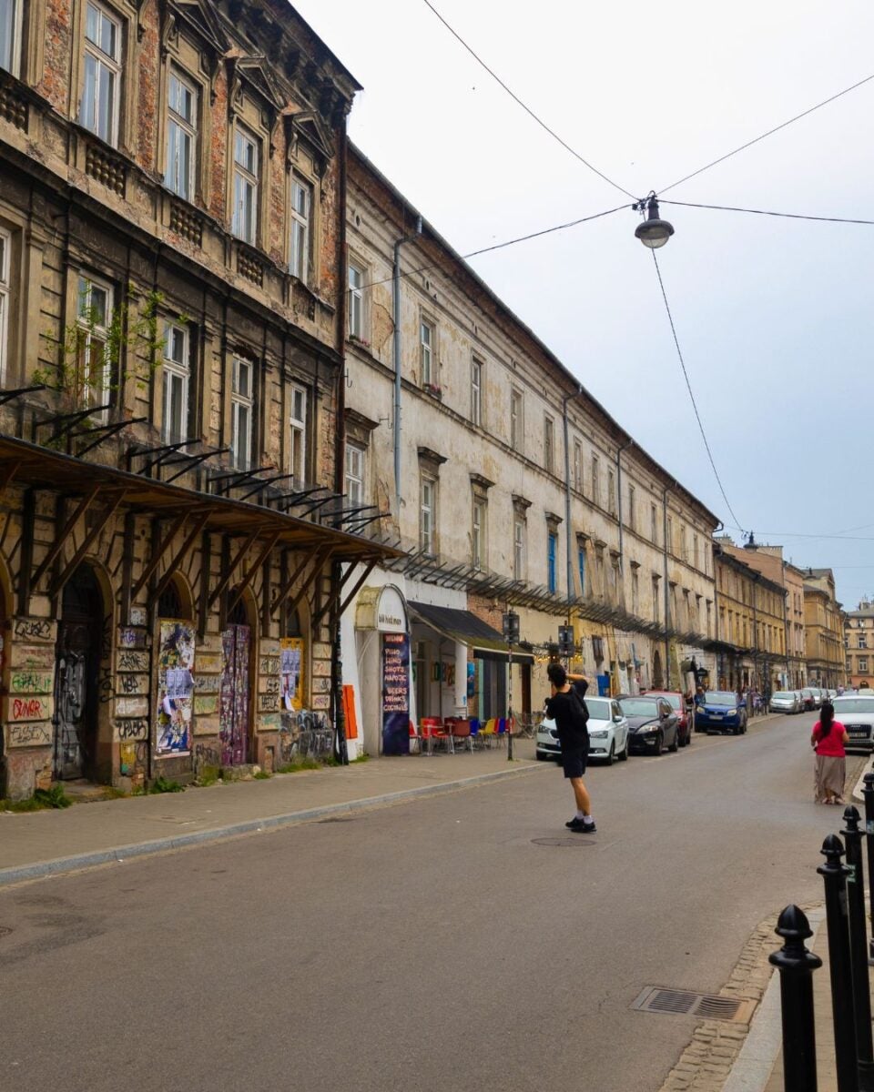 Old European street with weathered buildings and a person walking in the middle of the road under a hanging streetlight.