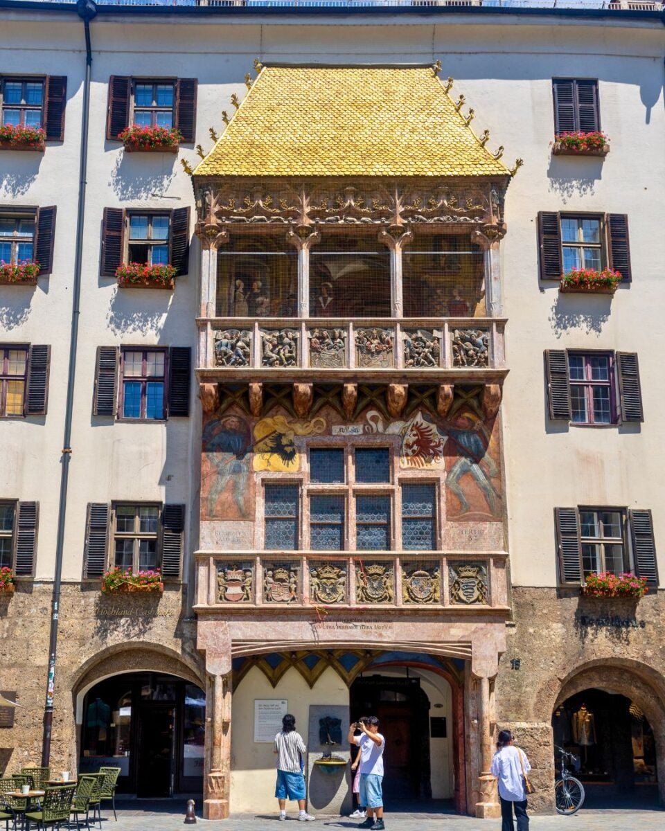 Historic building facade with a golden roof balcony and detailed frescoes above arched entrances in a European city.