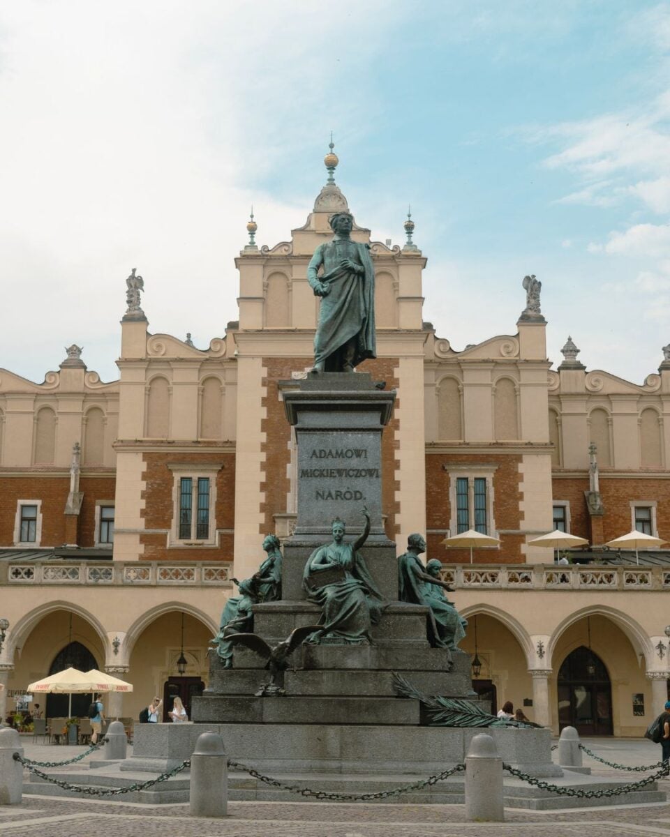 Statue of a historical figure atop a pedestal with four seated figures, set in front of an ornate building with arches and statues under a blue sky.