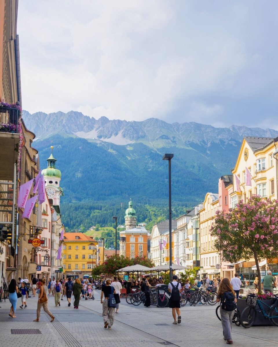 Bustling pedestrian street in a European town with colorful buildings and mountain range in the background under a cloudy sky.