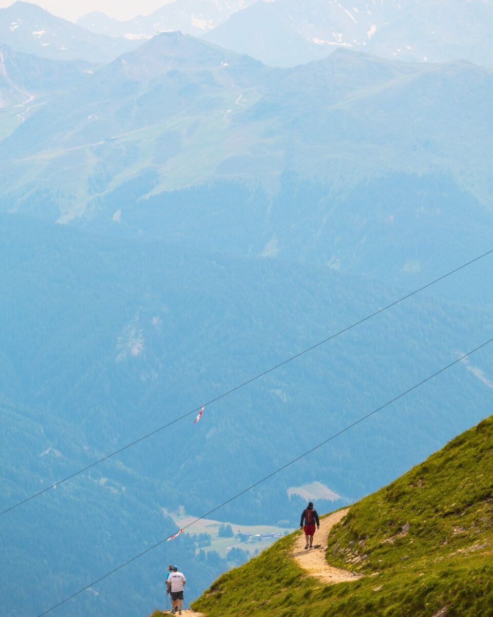 Hiker walking along a narrow mountain trail with another person further ahead, surrounded by vast green slopes and distant peaks.