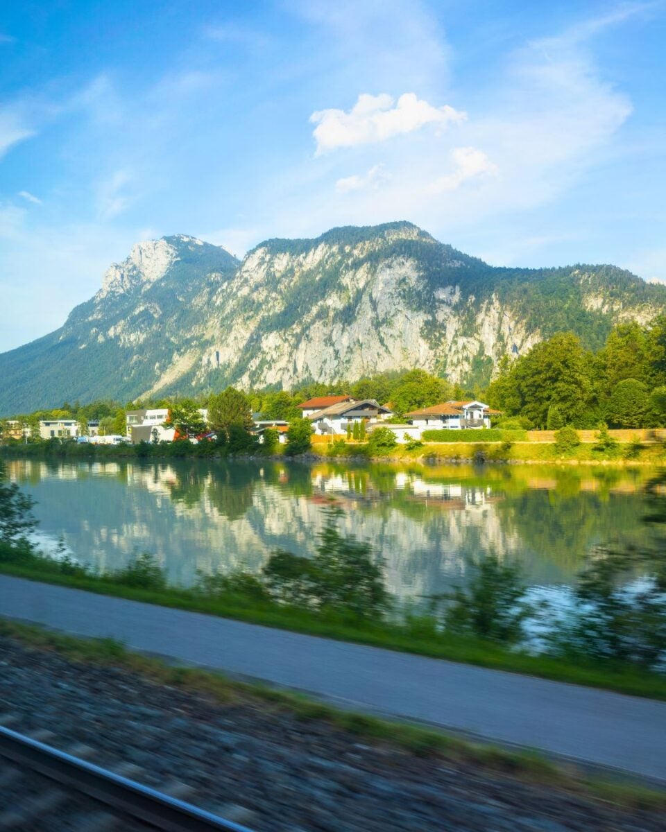 Scenic view of a mountain range reflected in a calm river with houses and greenery along the shore under a blue sky.