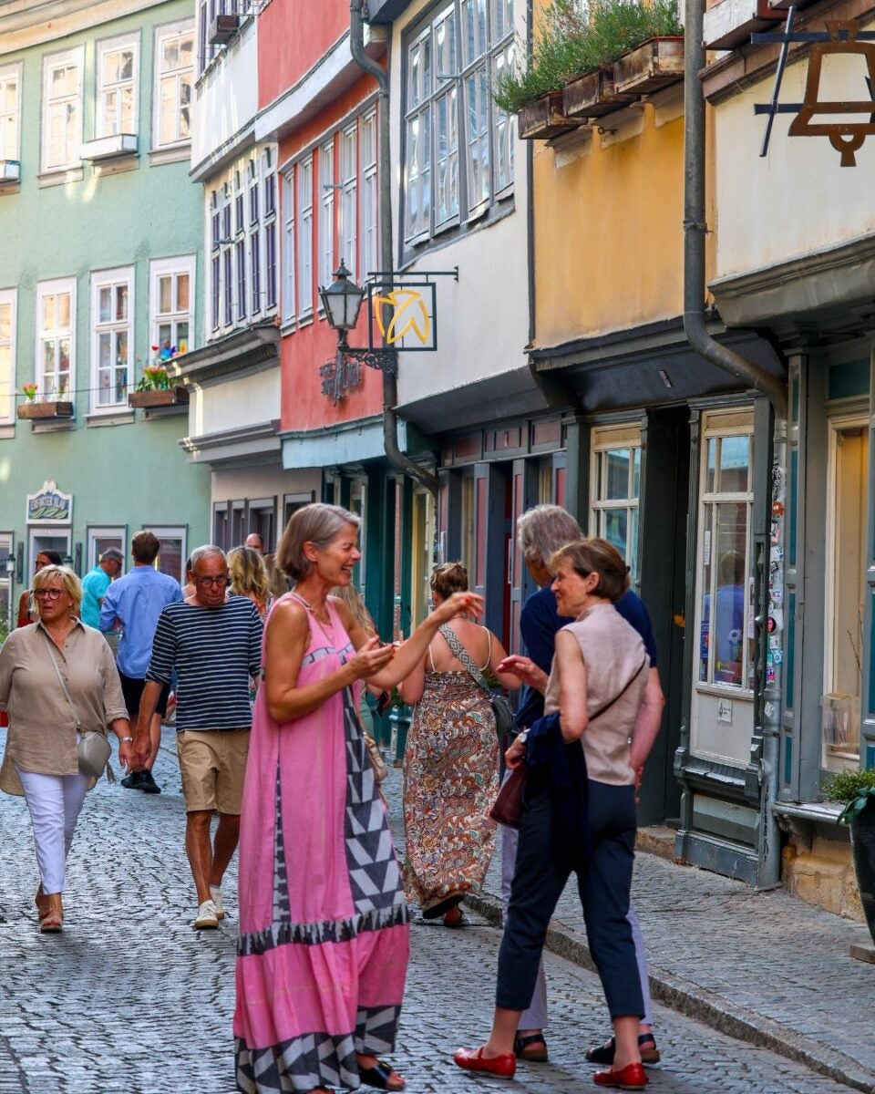People casually walking and chatting on a cobblestone street lined with colorful historic buildings and flower boxes.