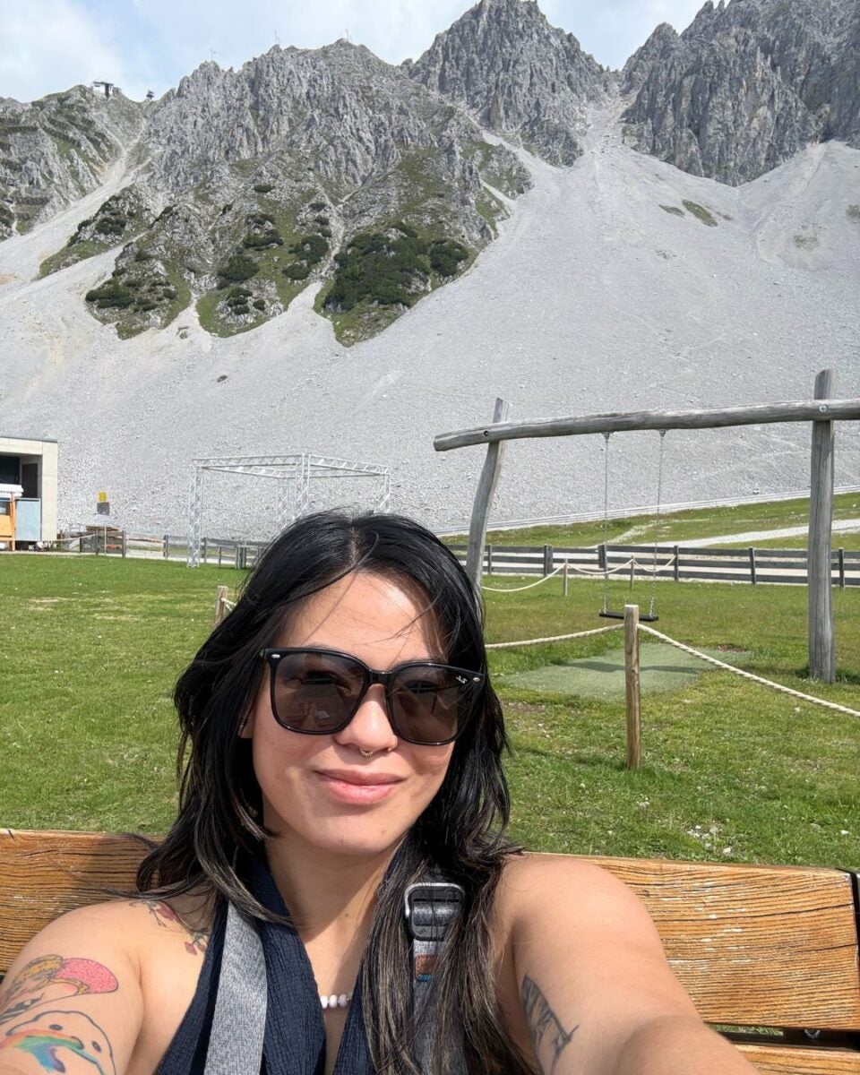 Woman with sunglasses and tattoos sitting on a wooden bench with rocky mountains and green grass behind her.