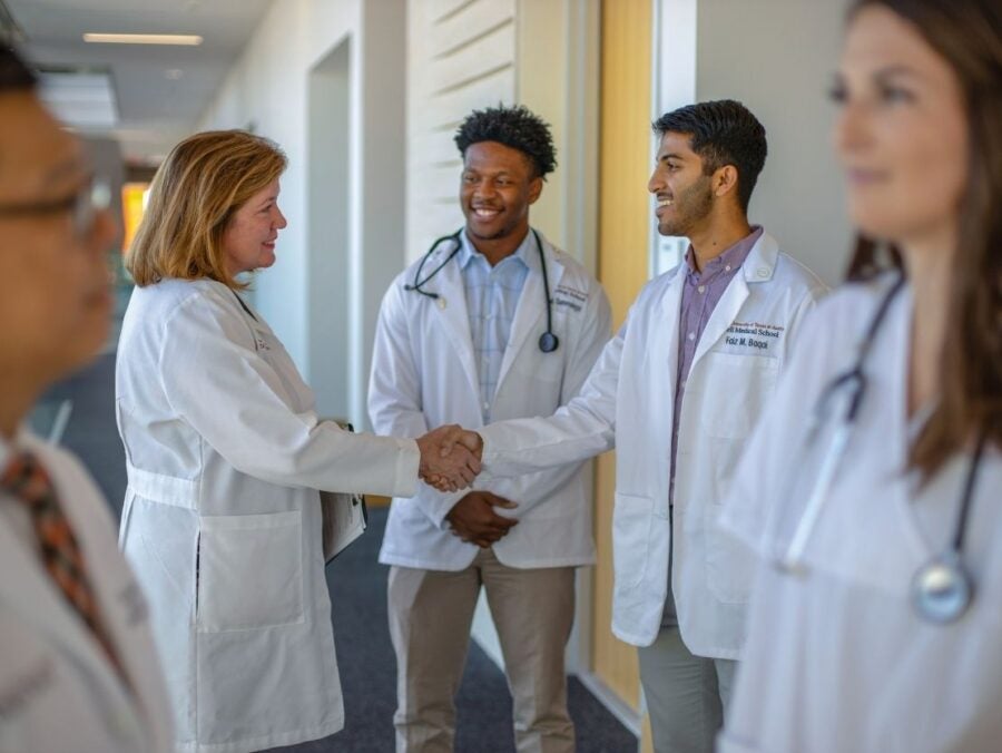 Doctors shaking hands in a hospital corridor while colleagues observe and smile nearby.