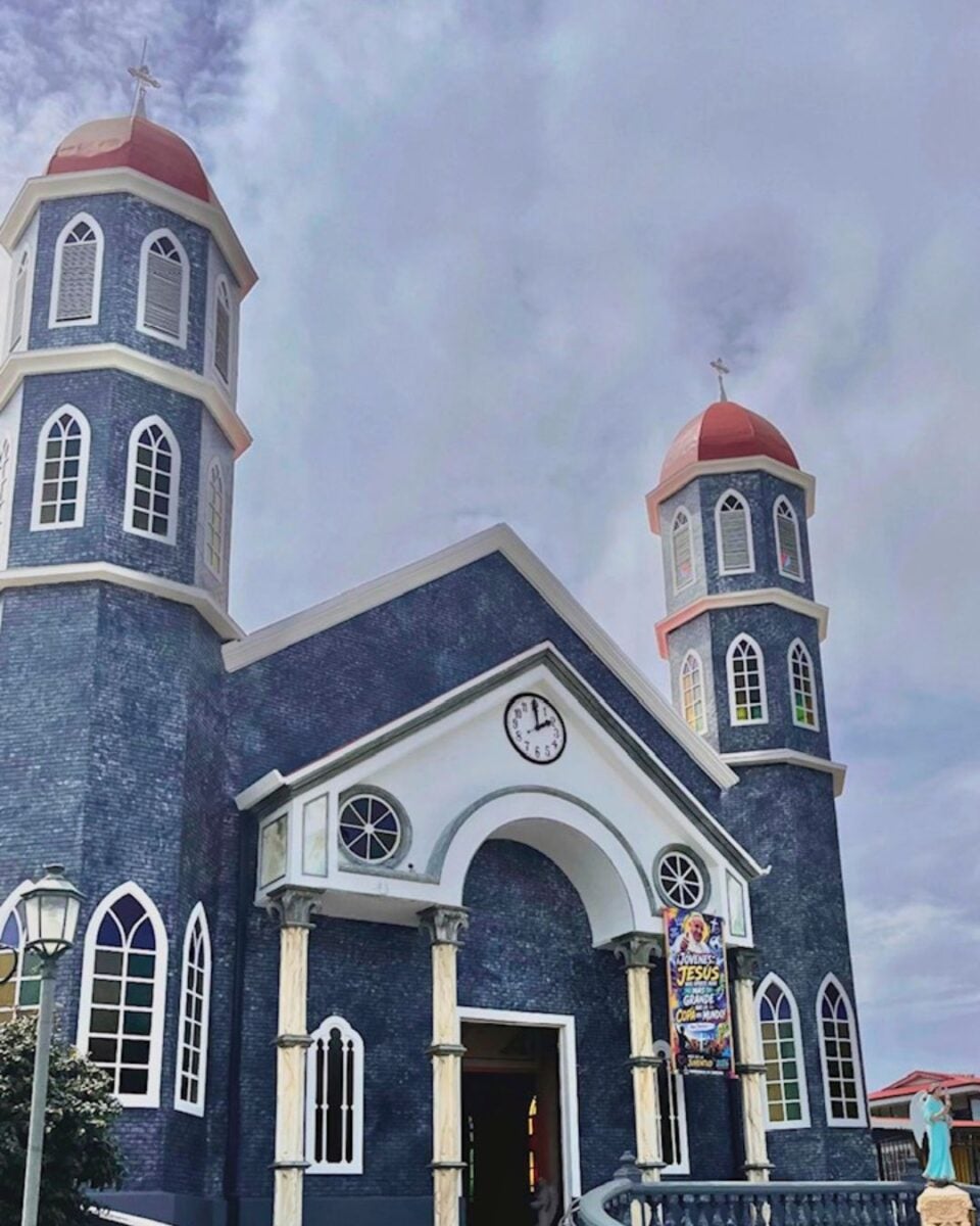 Blue church with two tall towers topped by red domes under a cloudy sky.
