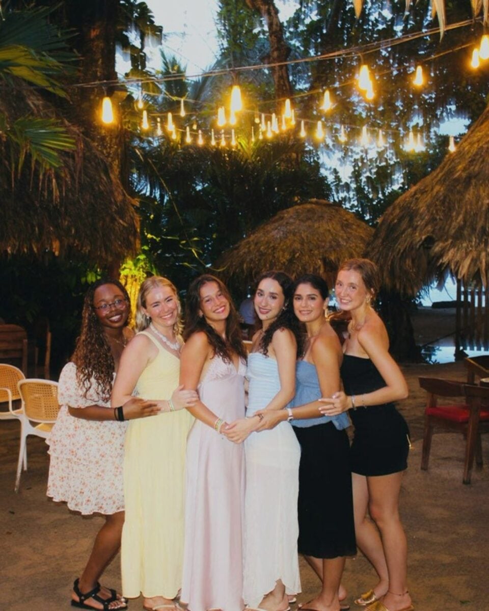 Six women in summer dresses smiling and posing together under string lights at an outdoor tropical venue at dusk.