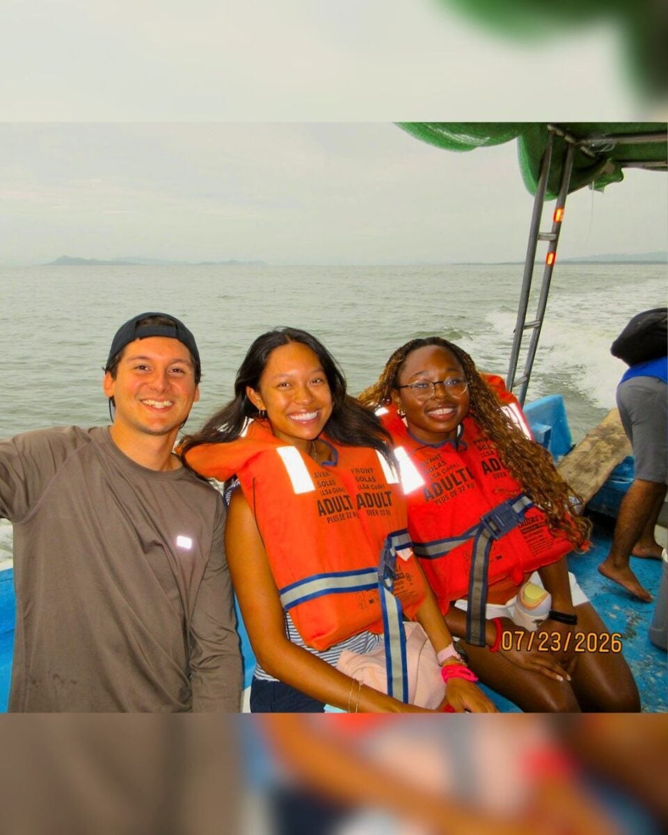 Three friends smiling on a boat wearing bright orange life jackets with calm water and distant land in the background.