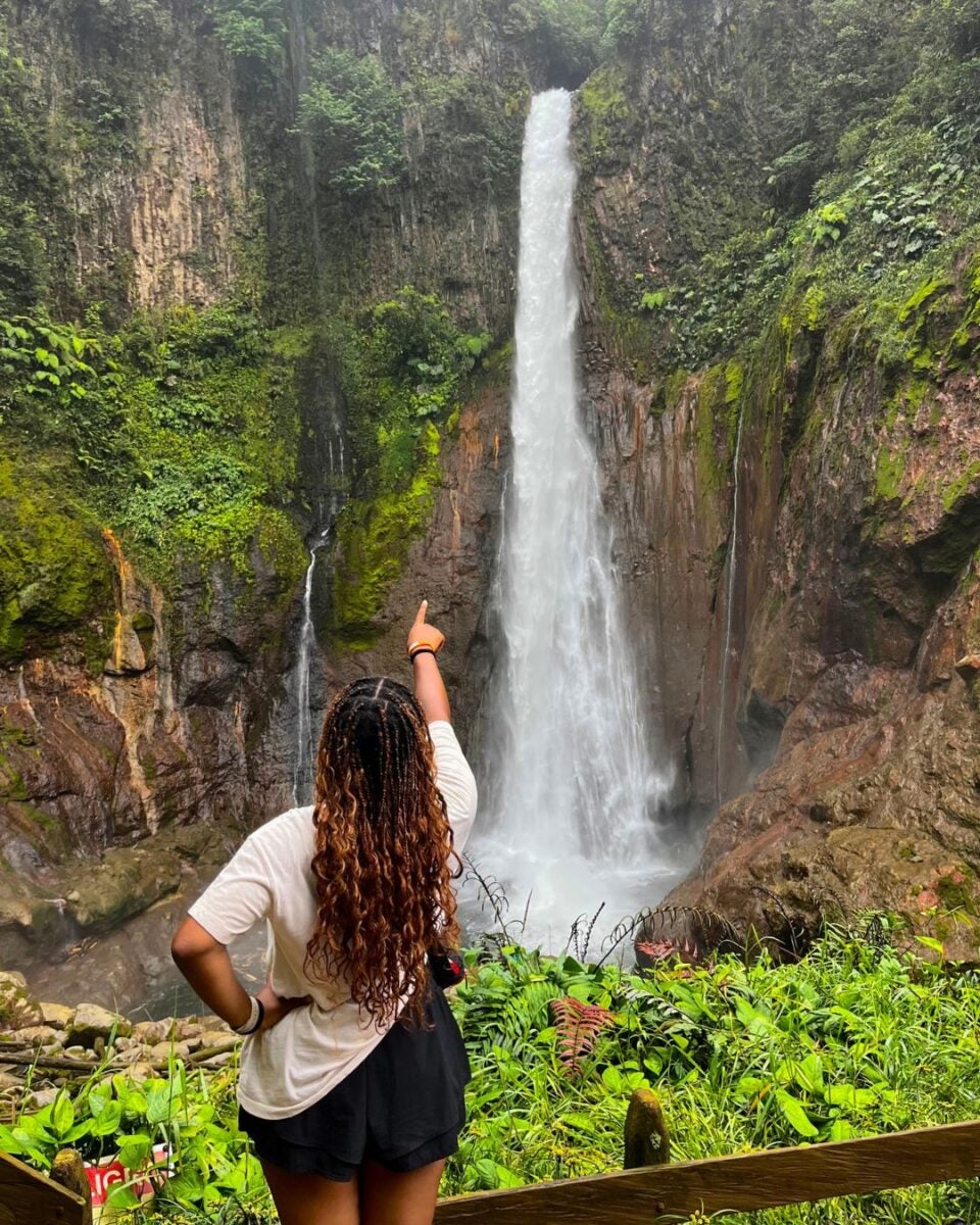 Woman with long curly hair points at a tall waterfall cascading down a rocky cliff surrounded by lush greenery.