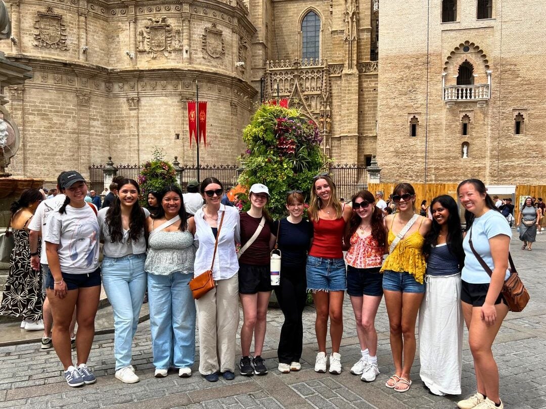Group of ten young women posing together in front of historic stone buildings on a sunny day in a European city square.