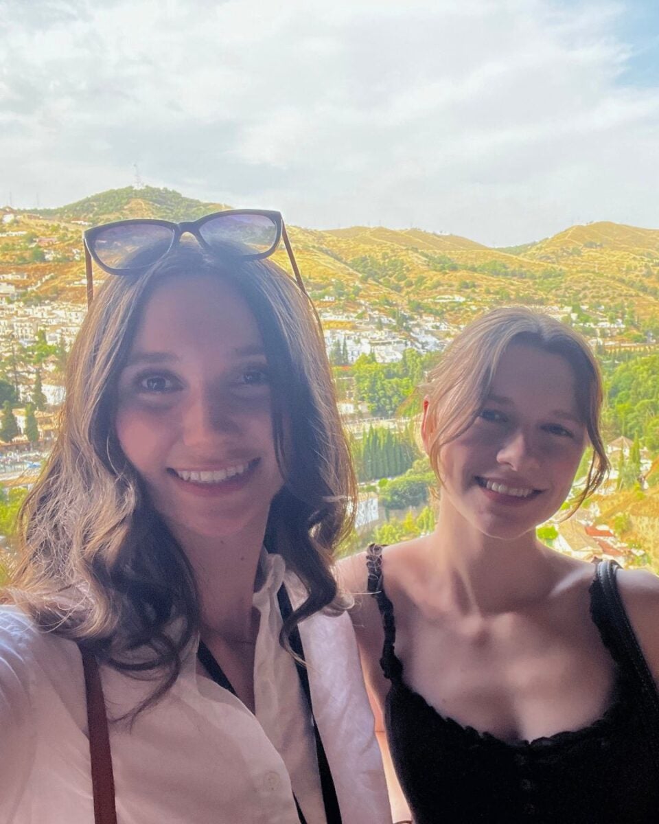 Two young women smiling for a selfie with a scenic hillside town and greenery in the background under a partly cloudy sky.
