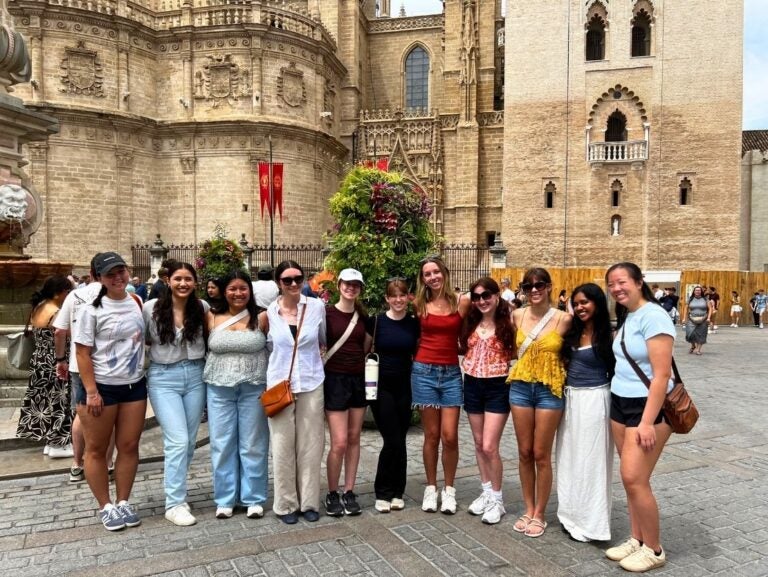 Group of ten young women posing together in front of historic stone buildings on a sunny day in a European city square.