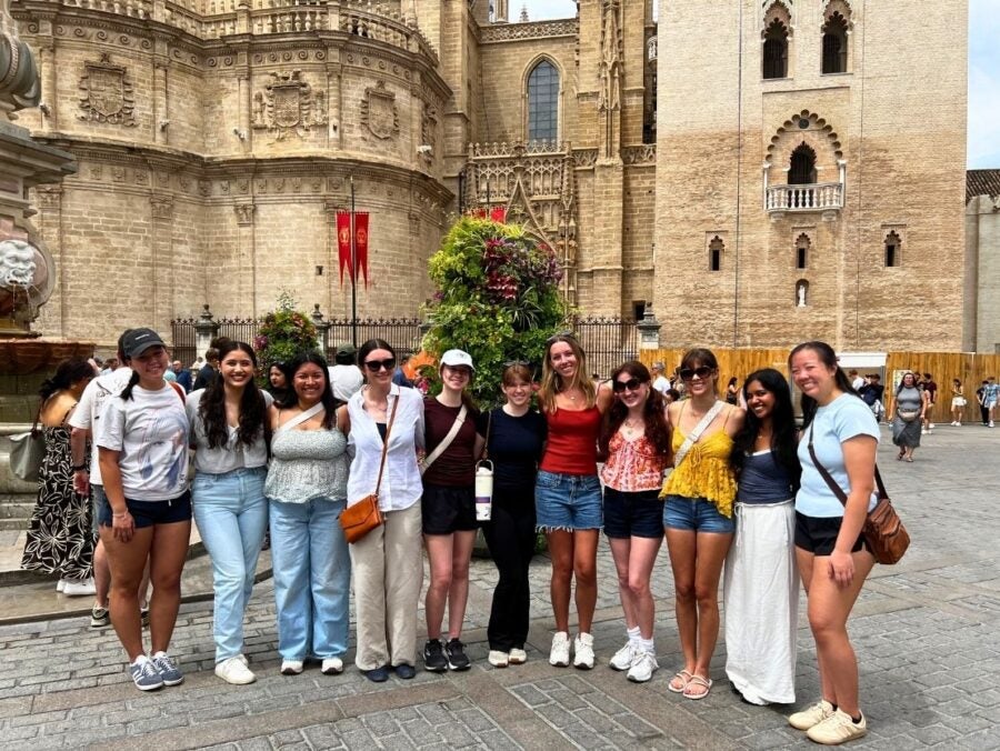 Group of ten young women posing together in front of historic stone buildings on a sunny day in a European city square.