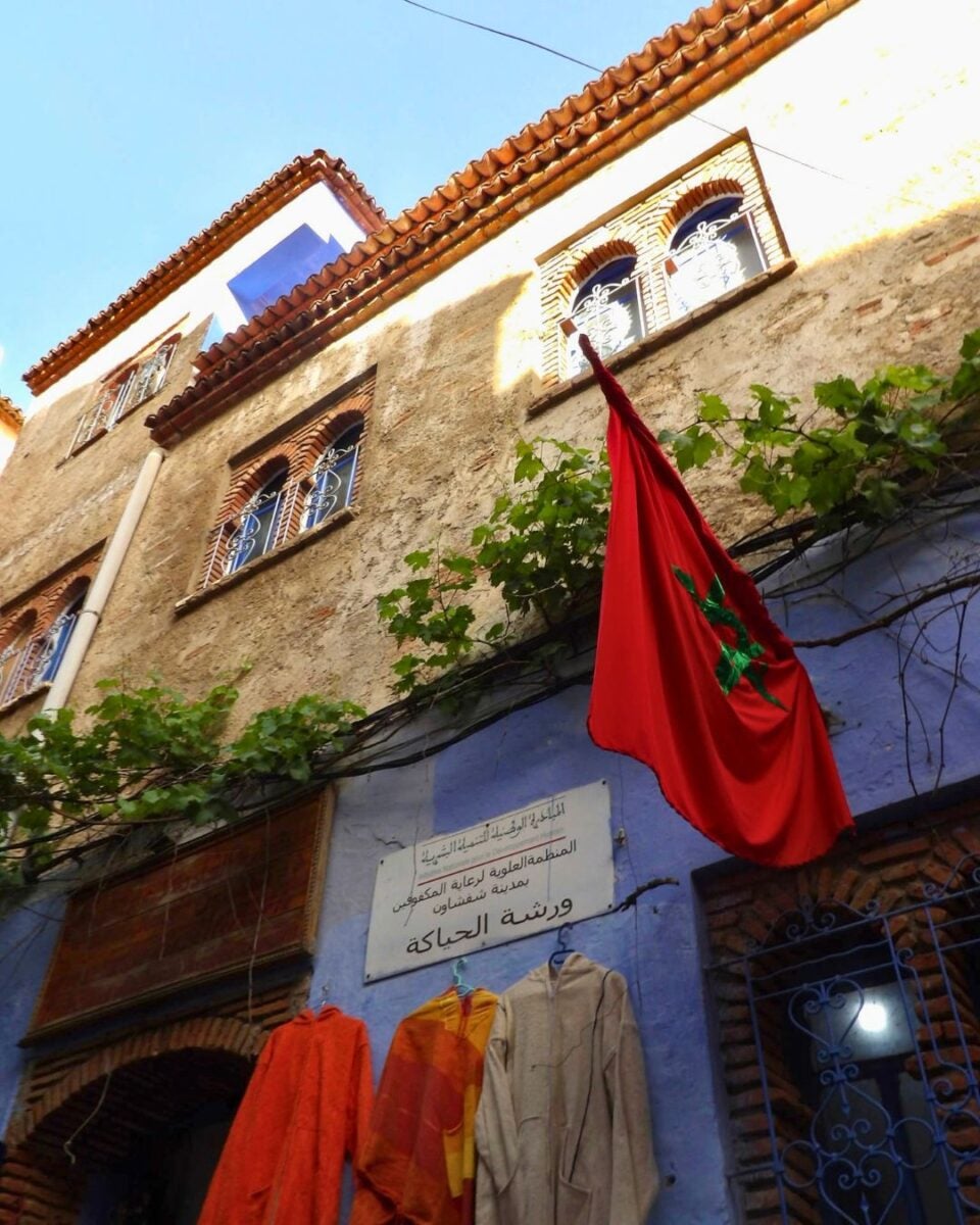 Moroccan flag hanging outside a traditional building with colorful robes displayed below under a clear blue sky.