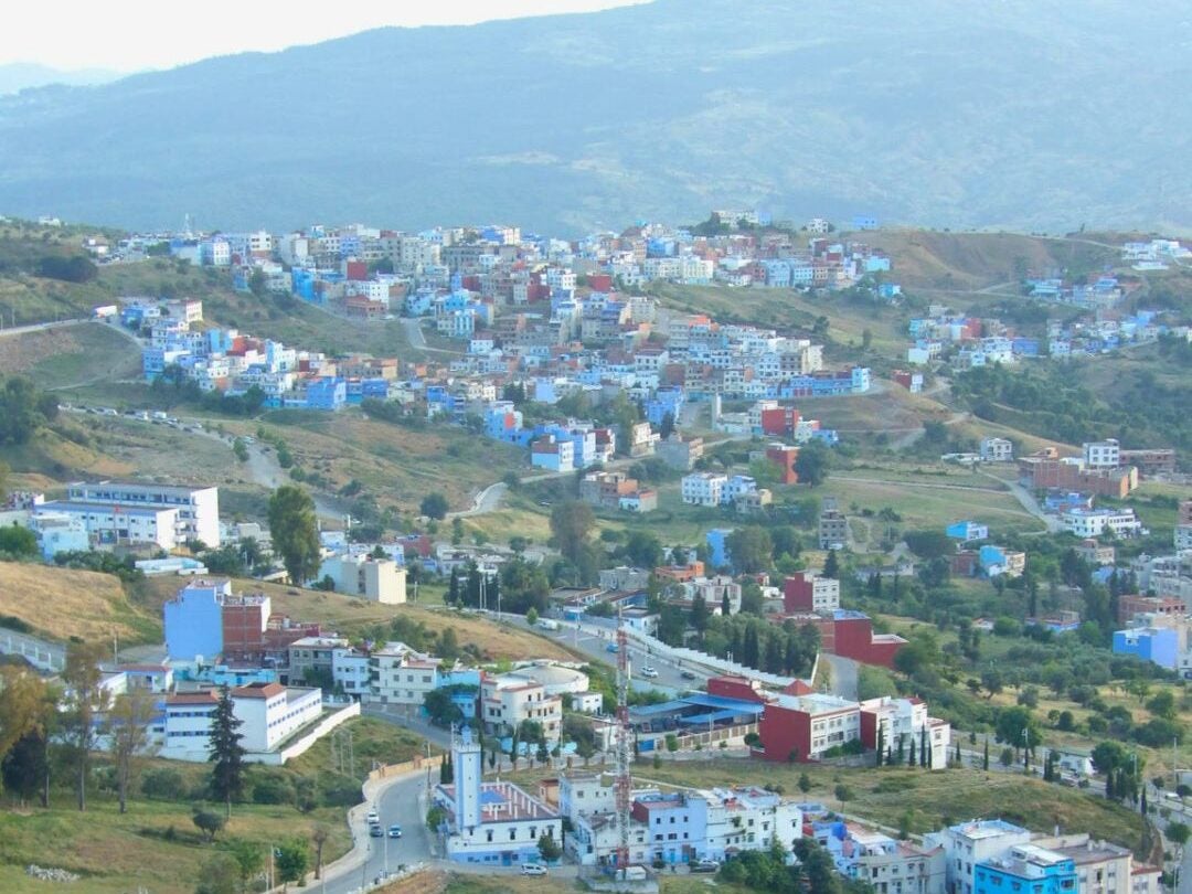 A panoramic view of a hillside town with colorful buildings scattered across green terrain under a hazy mountain backdrop.