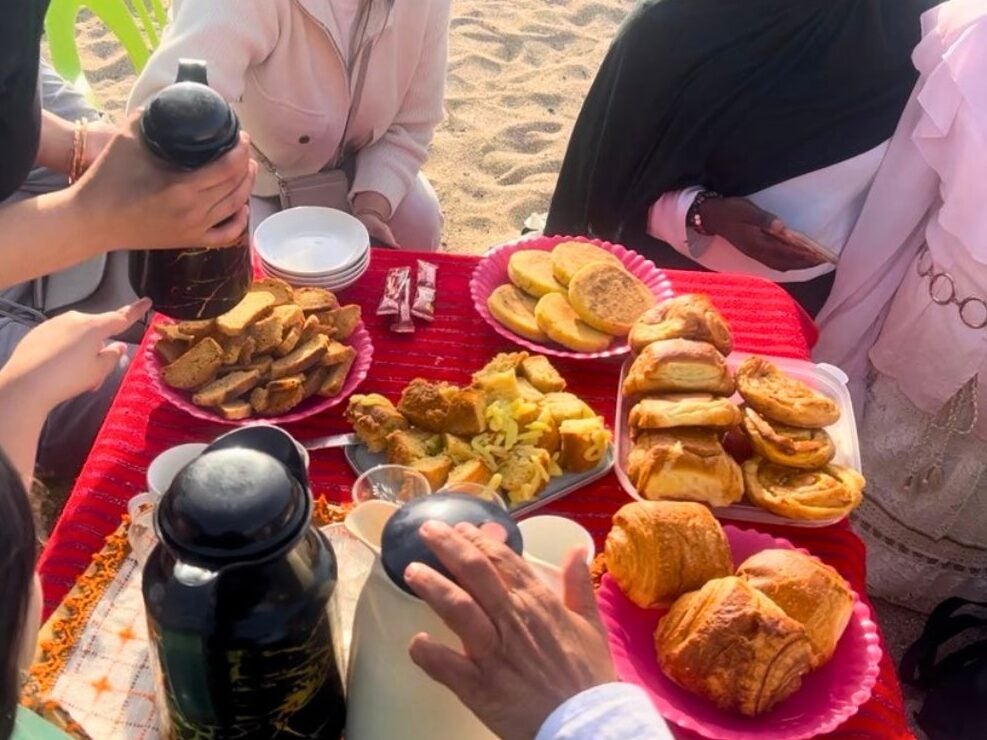 Group of people sharing a picnic with assorted pastries, snacks, and thermoses on a red tablecloth outdoors.