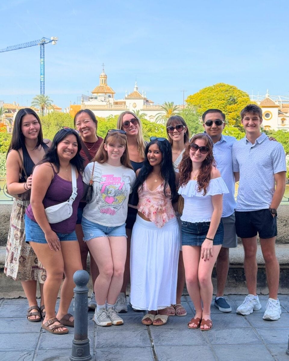 Group of nine young adults smiling outdoors with historic buildings and a crane in the background under a clear blue sky.