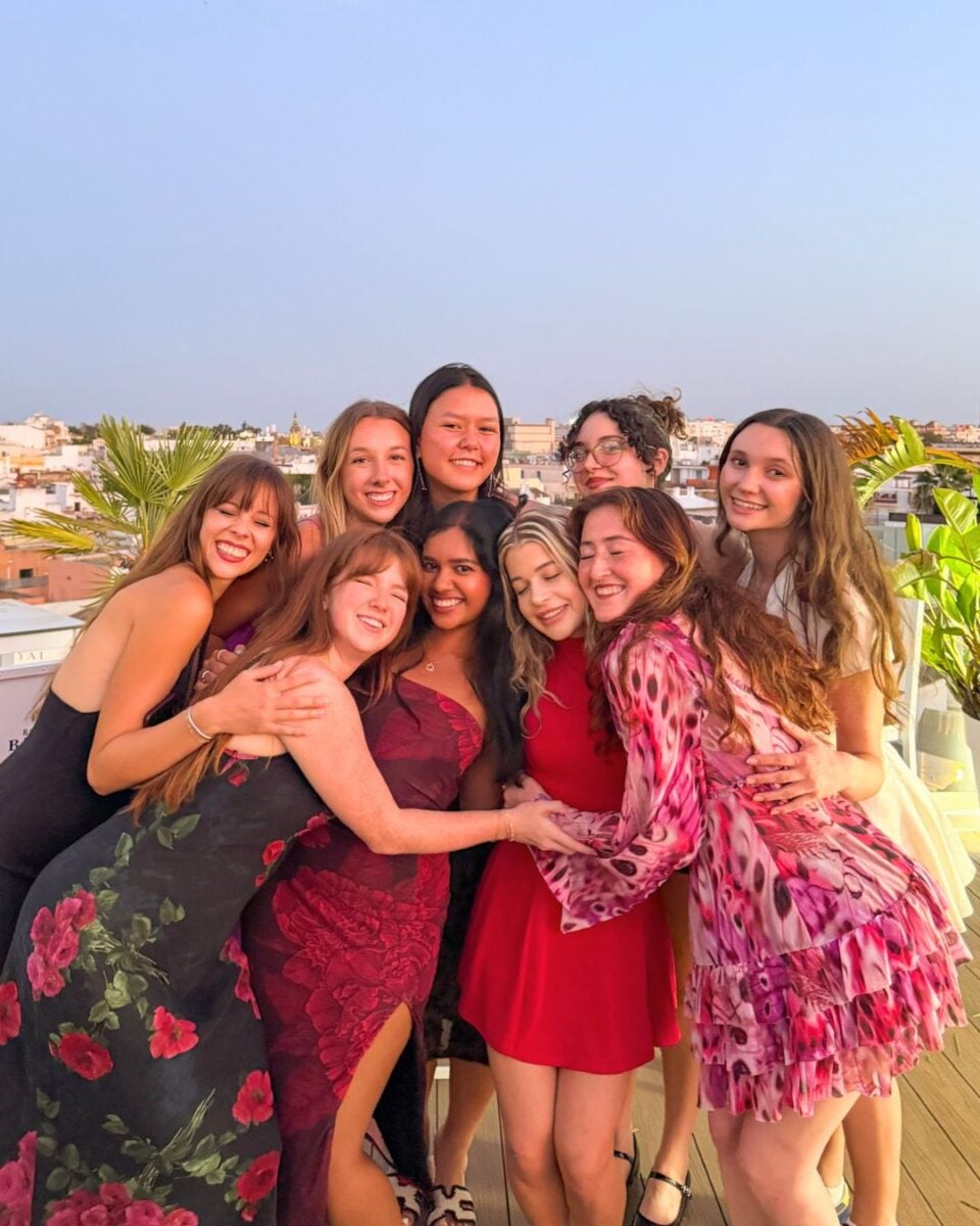 Group of eight young women in colorful dresses smiling and hugging on a rooftop with cityscape background at sunset.