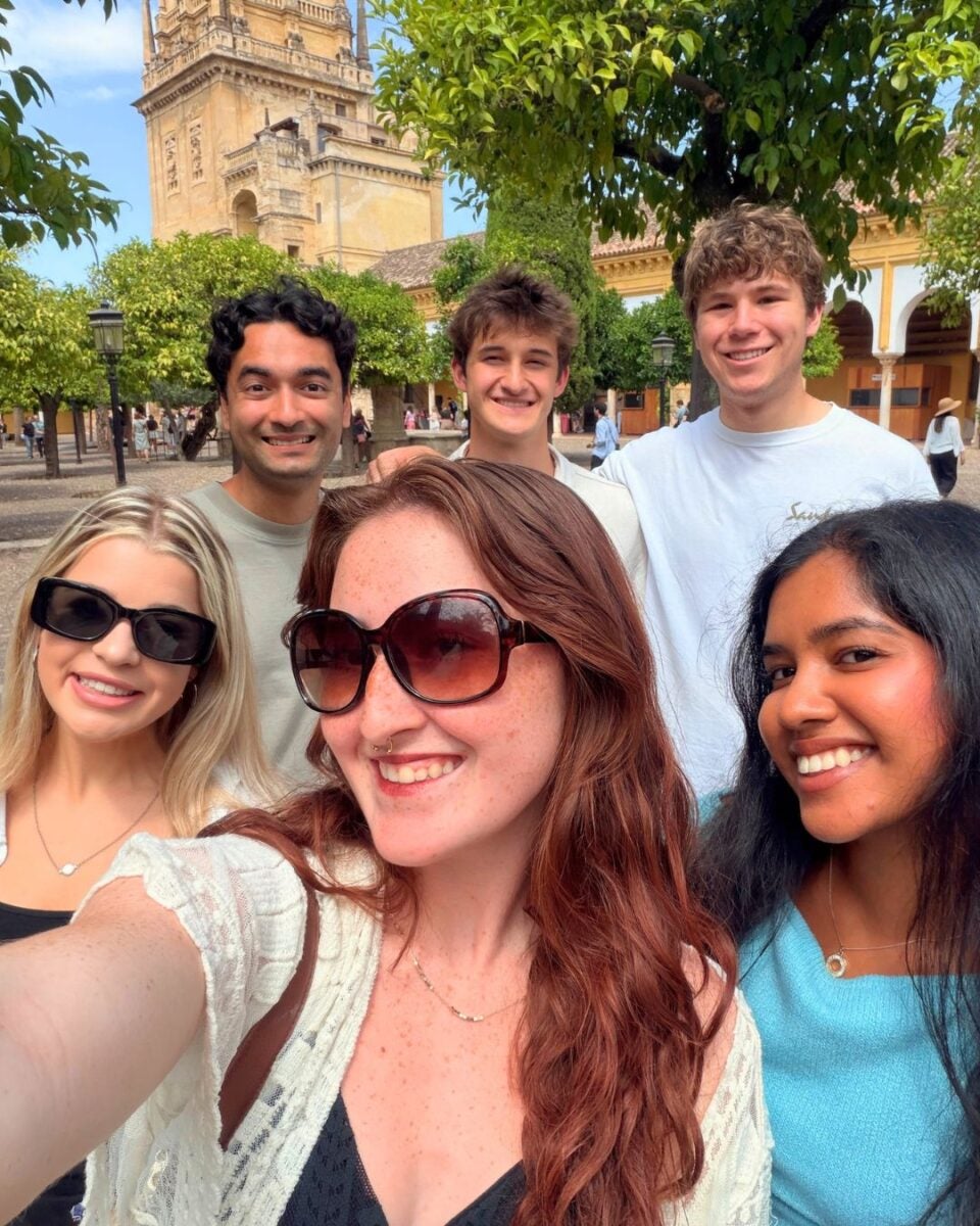 Group of six diverse friends smiling and taking a selfie outdoors near historic architecture and lush greenery on a sunny day.