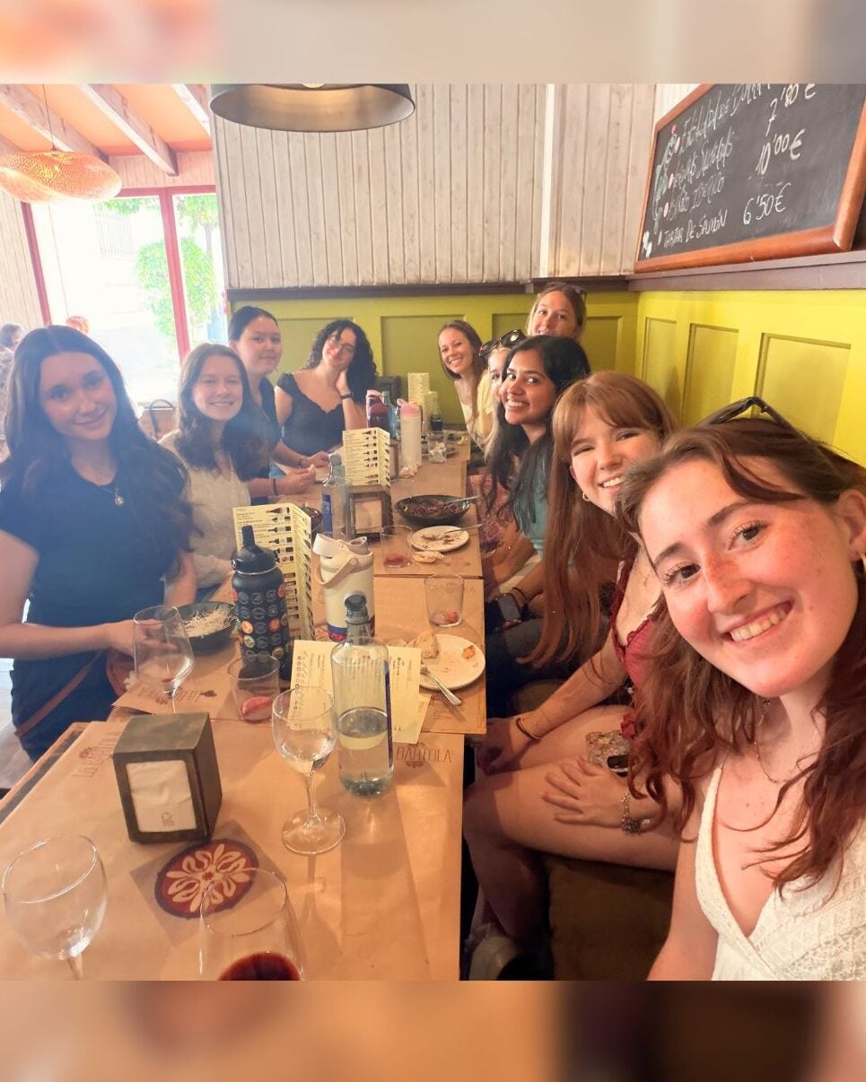 Group of nine young women smiling and enjoying a meal together at a cozy restaurant table with wooden decor and a chalkboard menu.