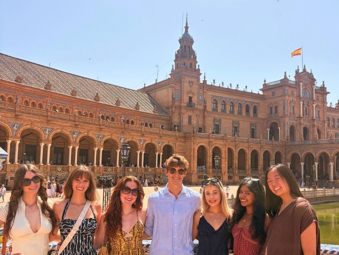 Group of six friends posing on a bridge at Plaza de España in Seville under clear blue sky.