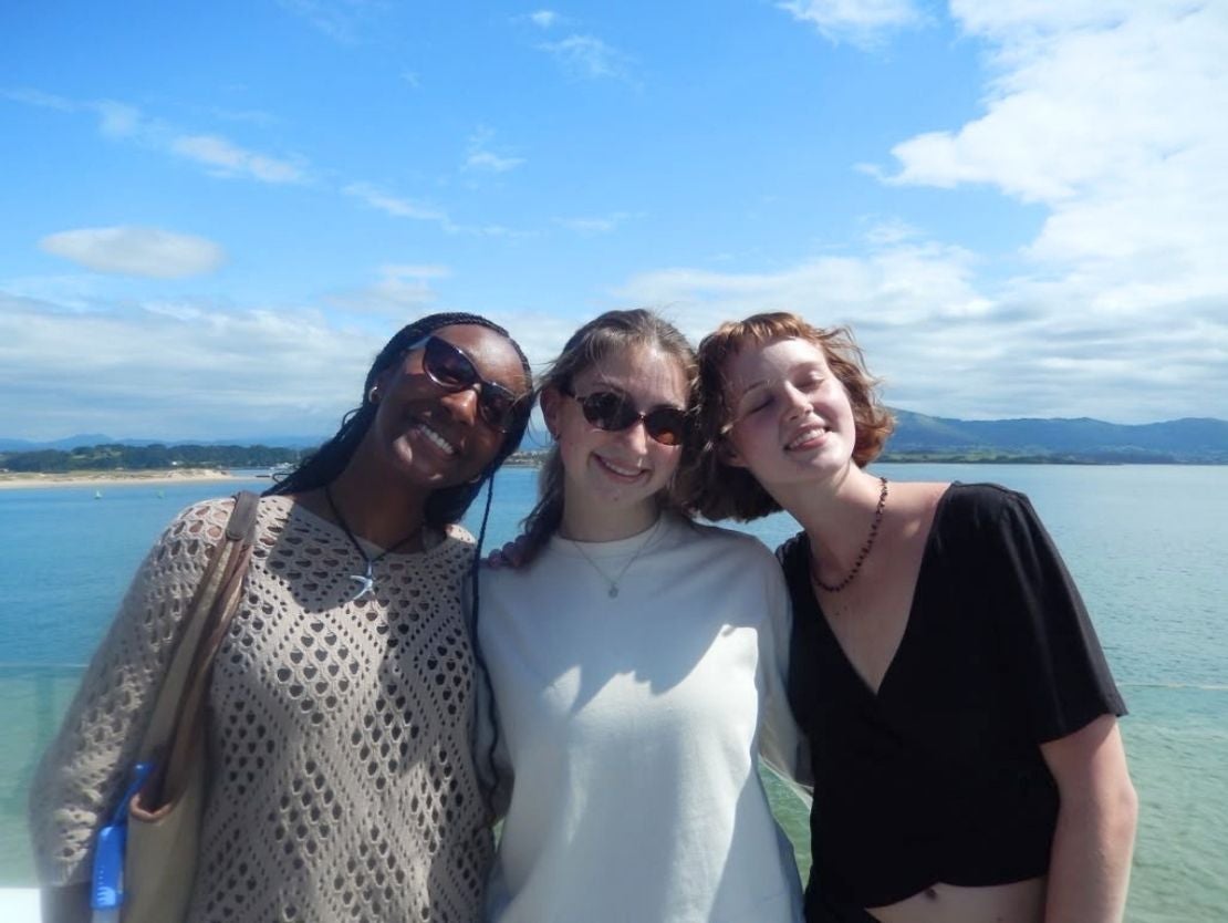 Three friends smiling and posing together by a sunny seaside with blue skies and distant hills.