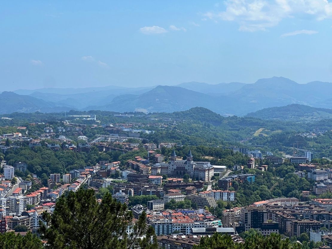A panoramic view of a city nestled among green hills with mountains in the distant background under a clear blue sky.