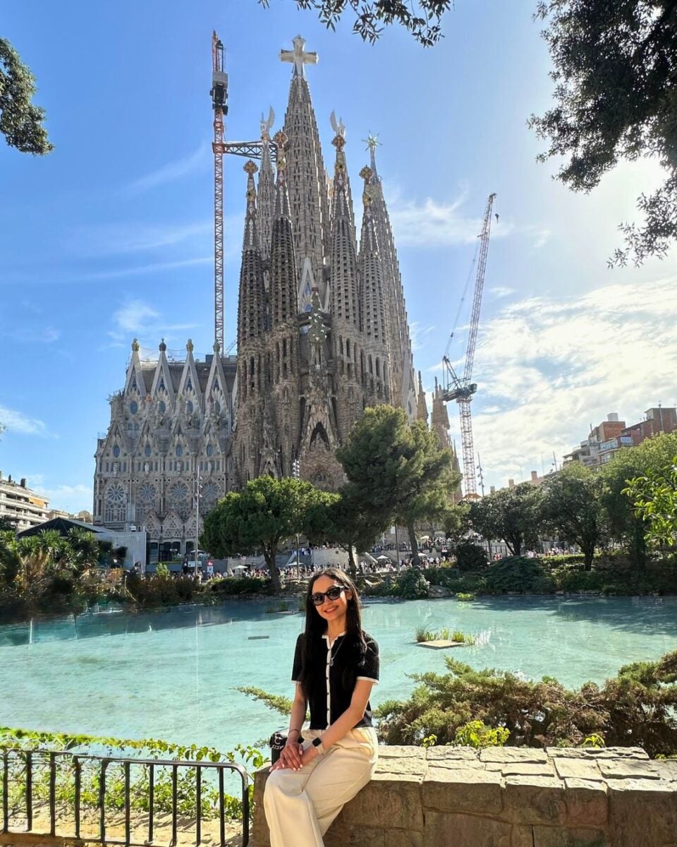 Woman in sunglasses sitting by a pond with the Sagrada Familia basilica and construction cranes in the background on a sunny day.
