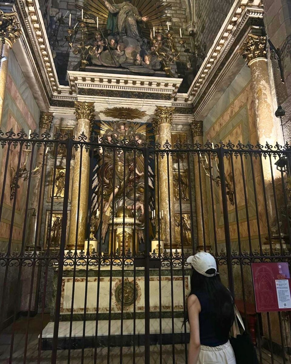 Woman in a white cap admiring a golden religious altar behind ornate iron gates inside a historic church.