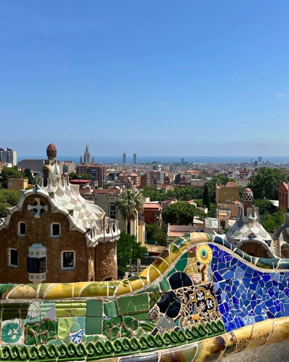 View of Barcelona cityscape from Park Güell with colorful mosaic benches and Gaudí's architectural buildings under a clear blue sky.