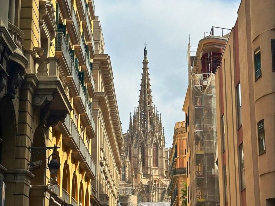 View of a Gothic cathedral spire rising above narrow city streets lined with historic buildings under a partly cloudy sky.