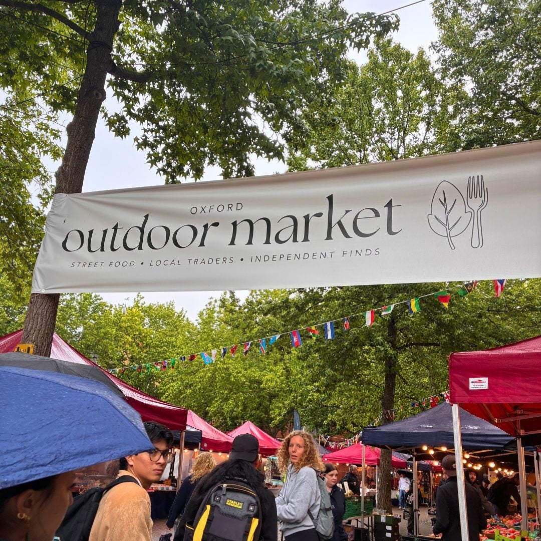 Crowded outdoor market in Oxford with red tents and shoppers browsing local vendors under a banner reading 'Oxford outdoor market'.