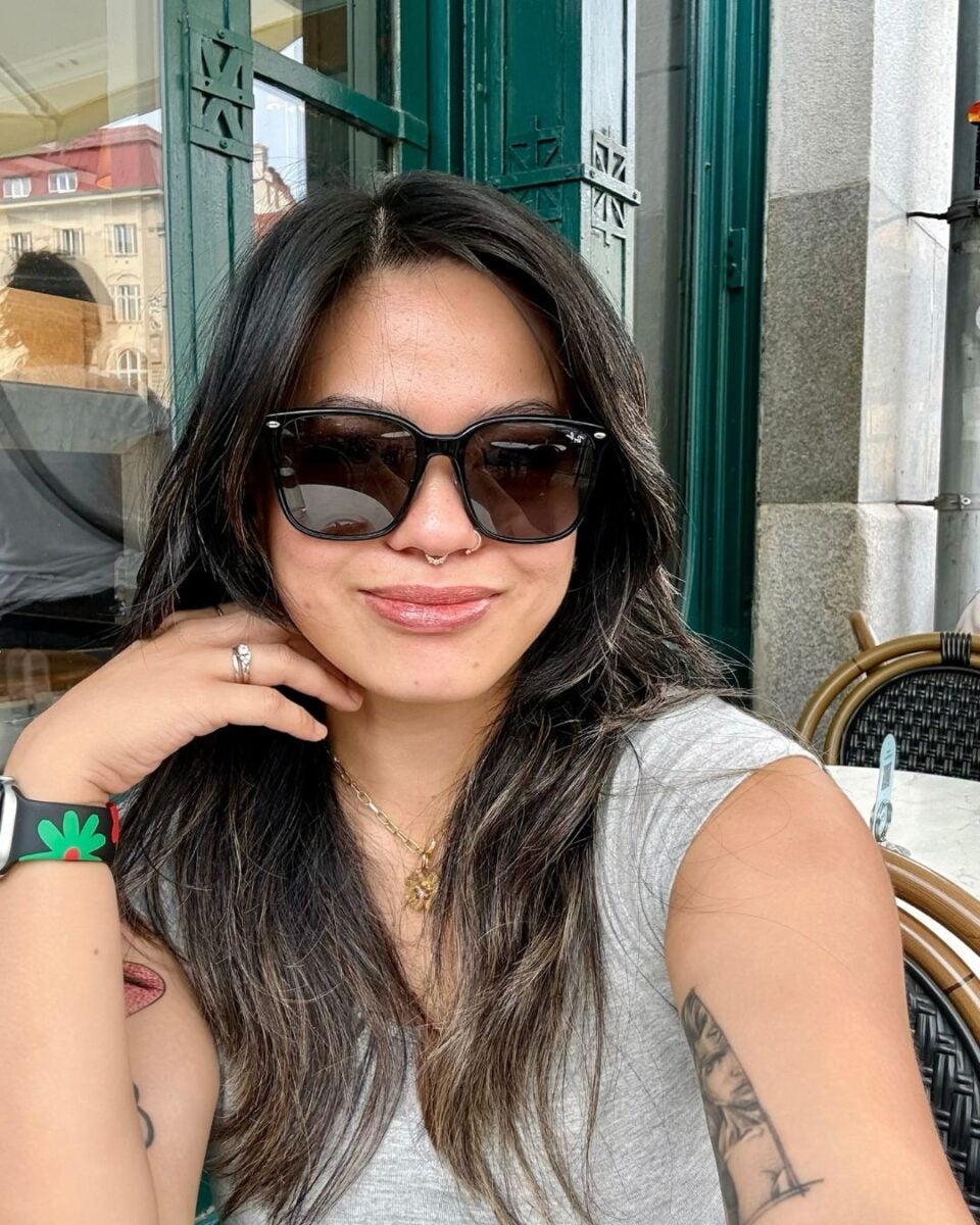 Young woman with long dark hair wearing large black sunglasses and a gray sleeveless top, sitting outdoors at a café with green window frames behind her.
