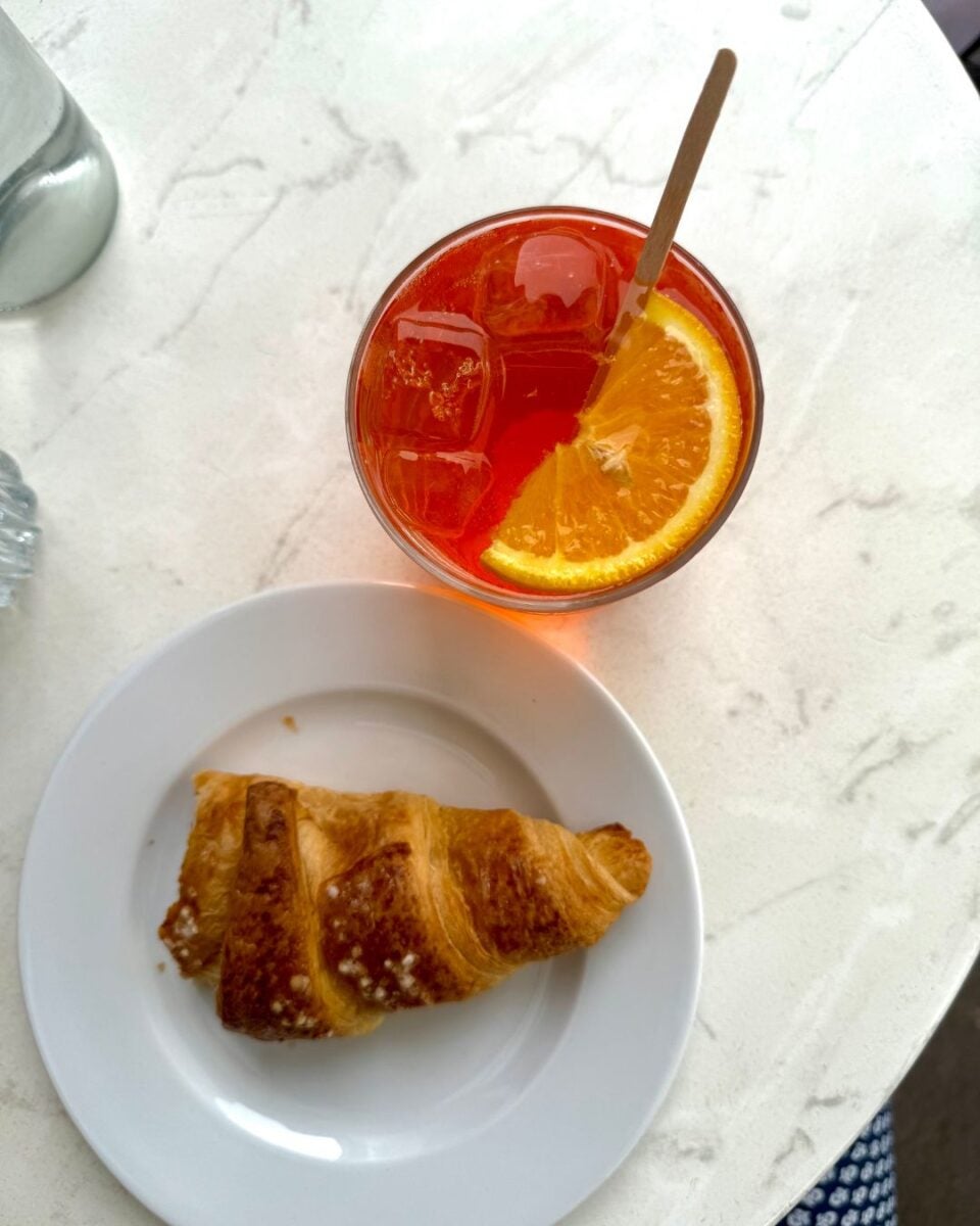 Top view of a croissant on a white plate beside a glass of iced orange cocktail with a slice of orange and a stirrer on a marble table.