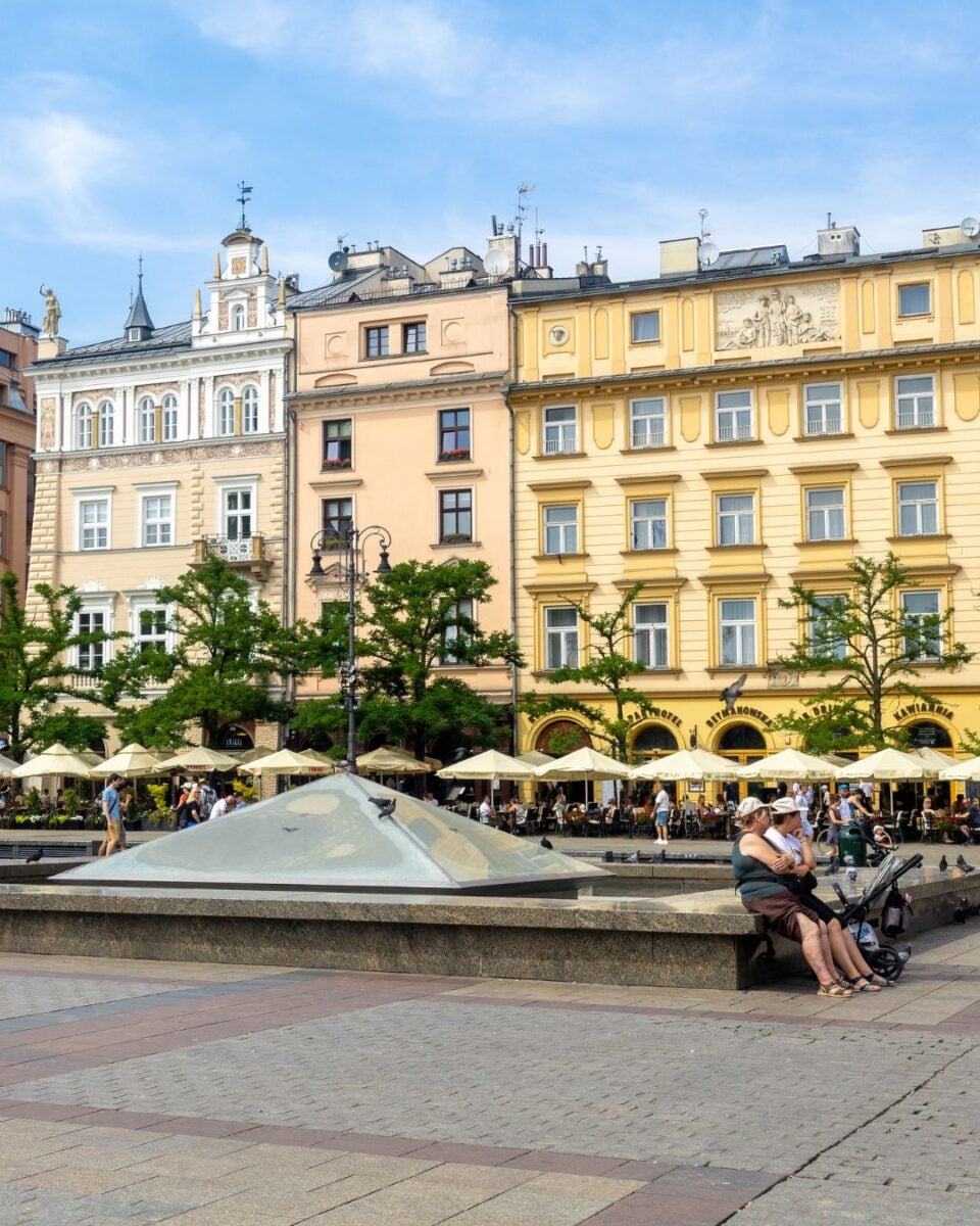 Historic European square with colorful buildings, outdoor cafes, and people relaxing near a modern fountain under a blue sky.