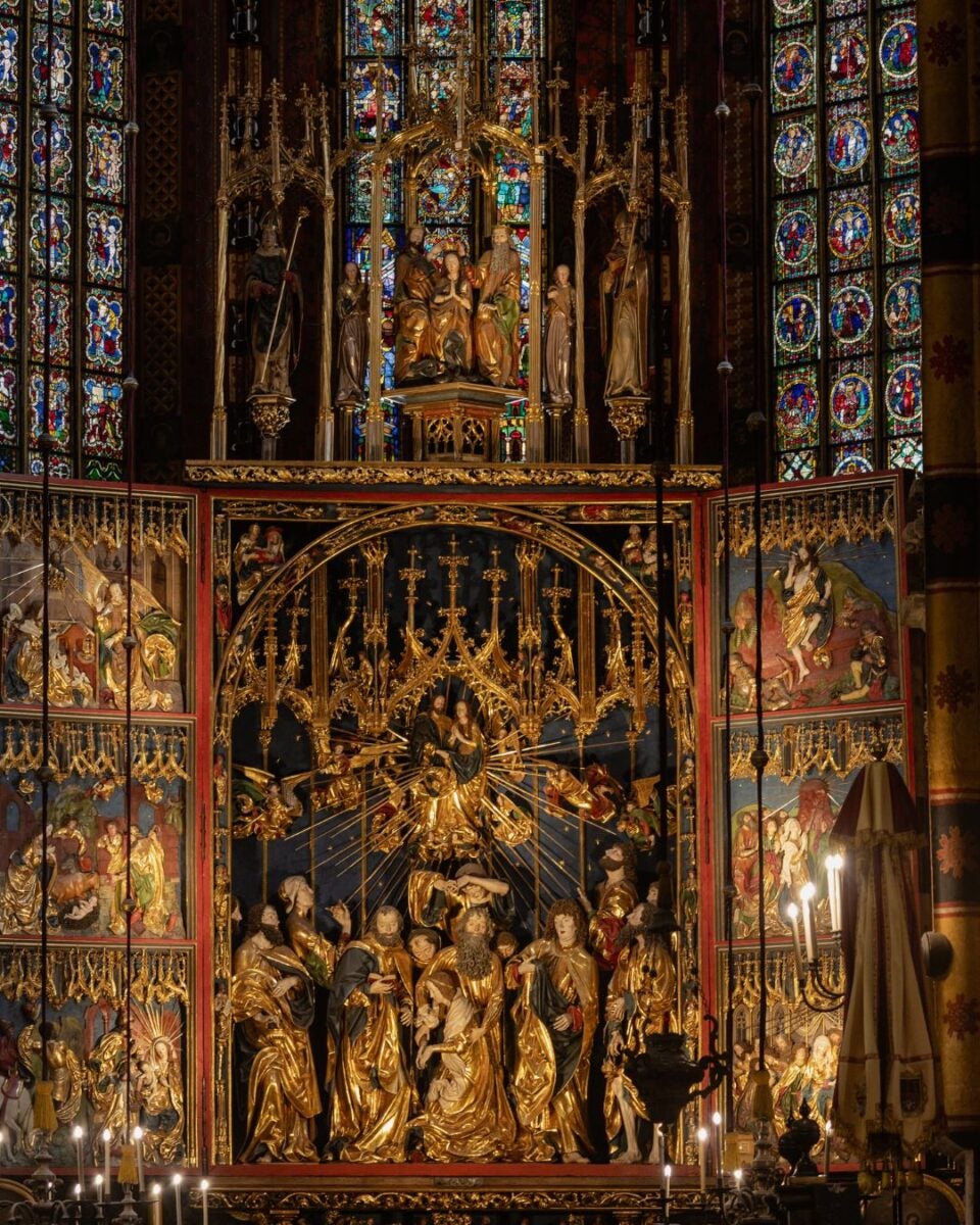 Intricately carved golden altar with religious figures and detailed stained glass windows in a dimly lit cathedral.