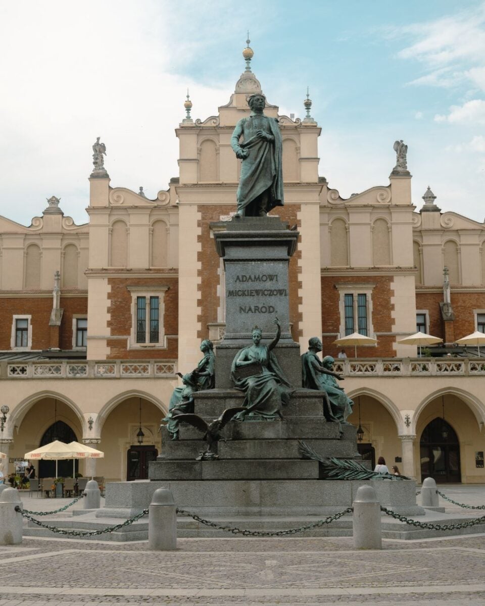 Bronze statue of Adam Mickiewicz atop a pedestal with allegorical figures, set in a historic square with arched buildings behind it.