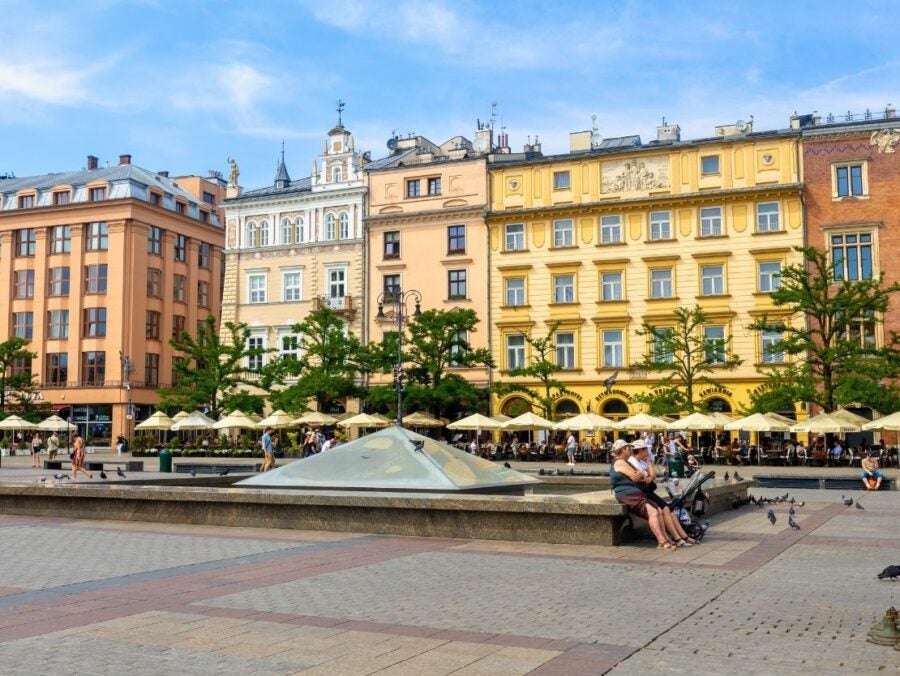 Historic European square with colorful buildings, outdoor cafes, and people relaxing near a modern fountain under a blue sky.