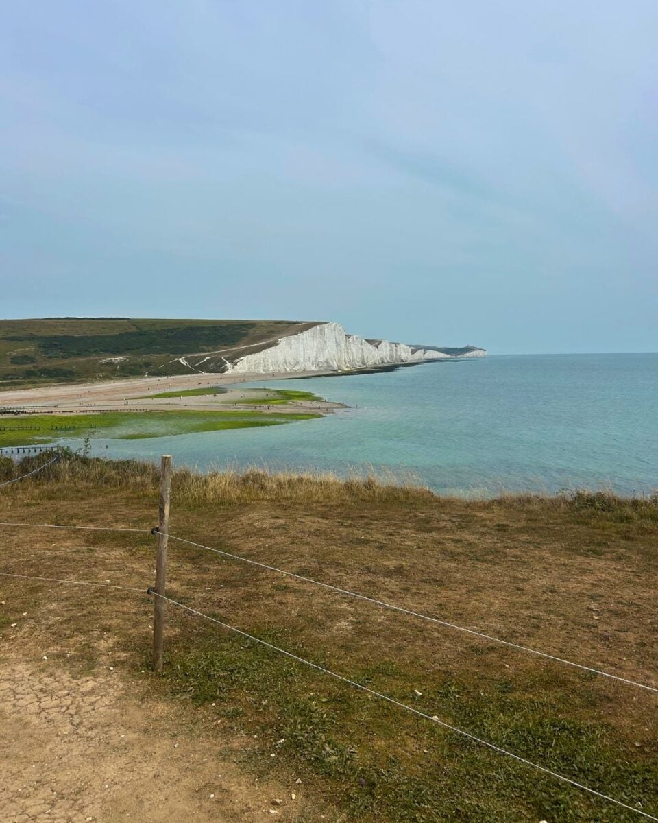 View of white chalk cliffs along a calm sea under a pale blue sky with grassy foreground and a wire fence.