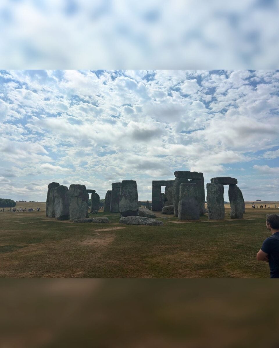 Wide view of Stonehenge's ancient stone circle under a partly cloudy sky on a grassy plain.