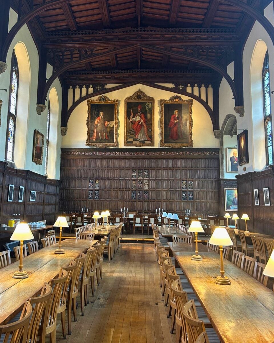 Historic dining hall with long wooden tables, chairs, and lit lamps under a vaulted wooden ceiling and large classical portraits on the far wall.